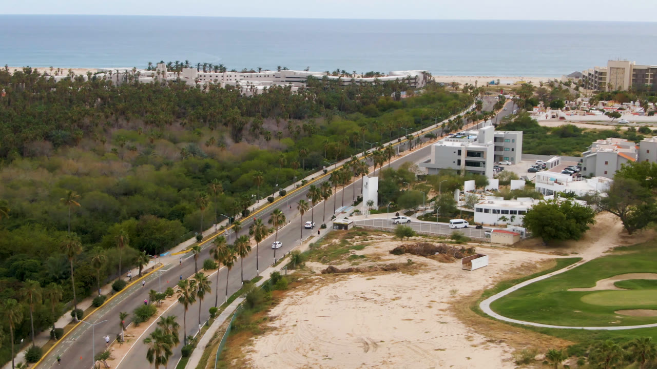 Drone frame of a coastal district with road, trees, buildings, sand, golf course, beach, and ocean.