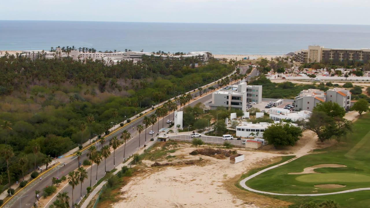 Opening aerial frame over San Jose del Cabo with a palm-lined road at left, sandy foreground, a golf green at right, resort buildings near the beach, ocean beyond, and no visible people.