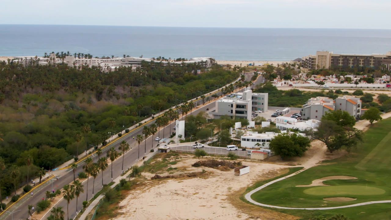 Aerial frame in San Jose del Cabo showing more of the golf fairway and sand traps on the right, white buildings near center, beach resort blocks near the ocean, and no visible people.