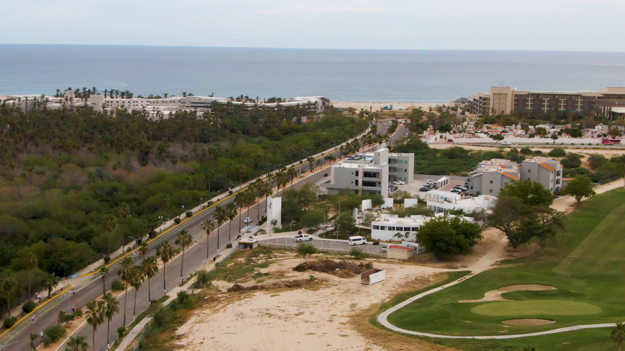 Aerial view over San Jose del Cabo with the road running diagonally along the left, low white buildings in the center, a curved golf green at lower right, beach resorts near the shore, and no visible people.