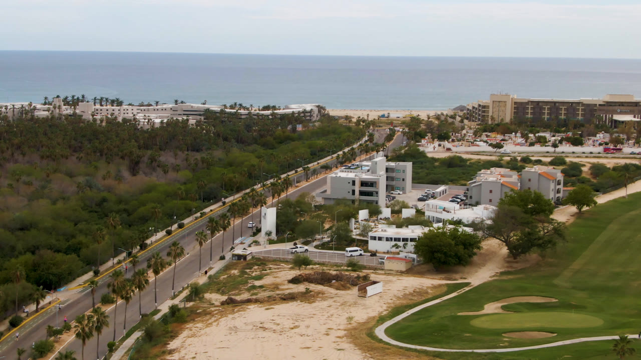 Aerial view of coastal trees, a palm-lined road, white buildings, golf course, resorts, and sea.