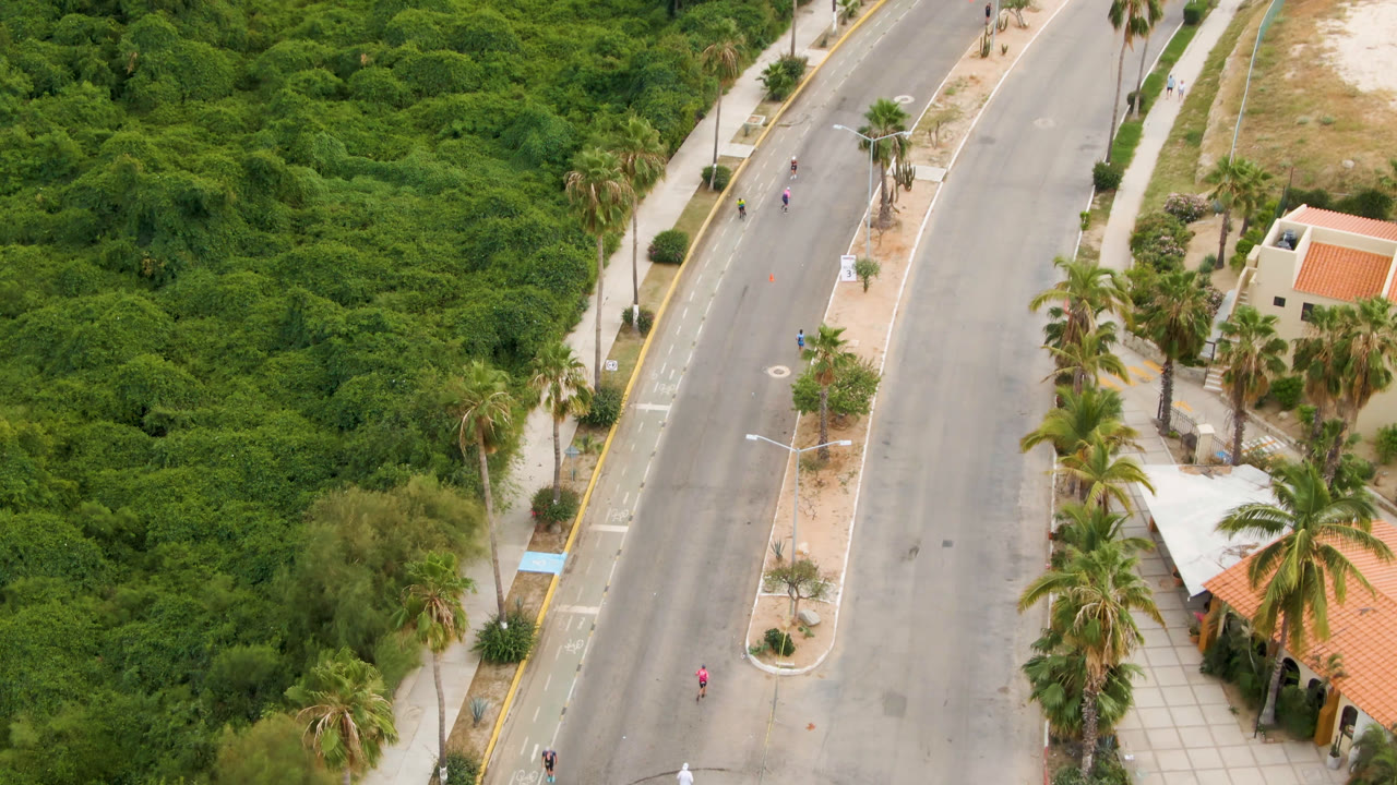 Second aerial frame in San Jose Del Cabo, Mexico, with runners farther along the paved road, greenery on one side and palm trees near small red-roofed buildings on the other.