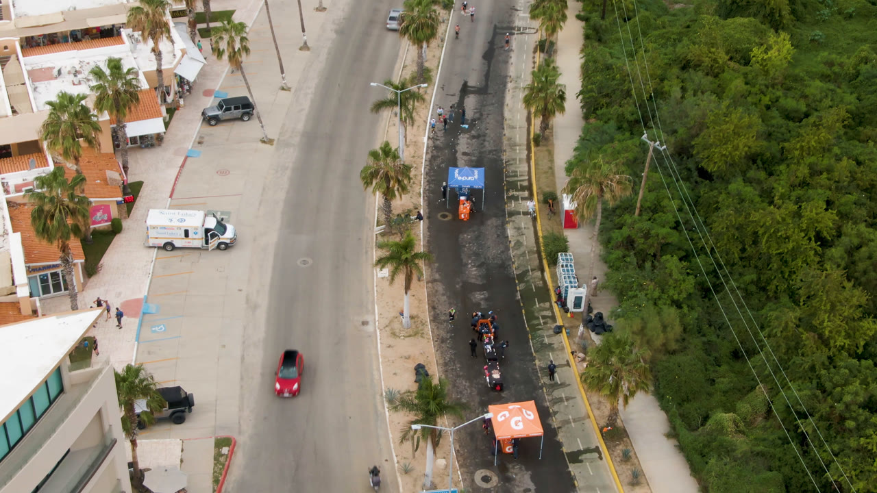Aerial view in San Jose del Cabo with a red car farther down the main road, a white truck by the building frontage, palms on both sides, a dark parallel lane, and small people visible near the sidewalk.