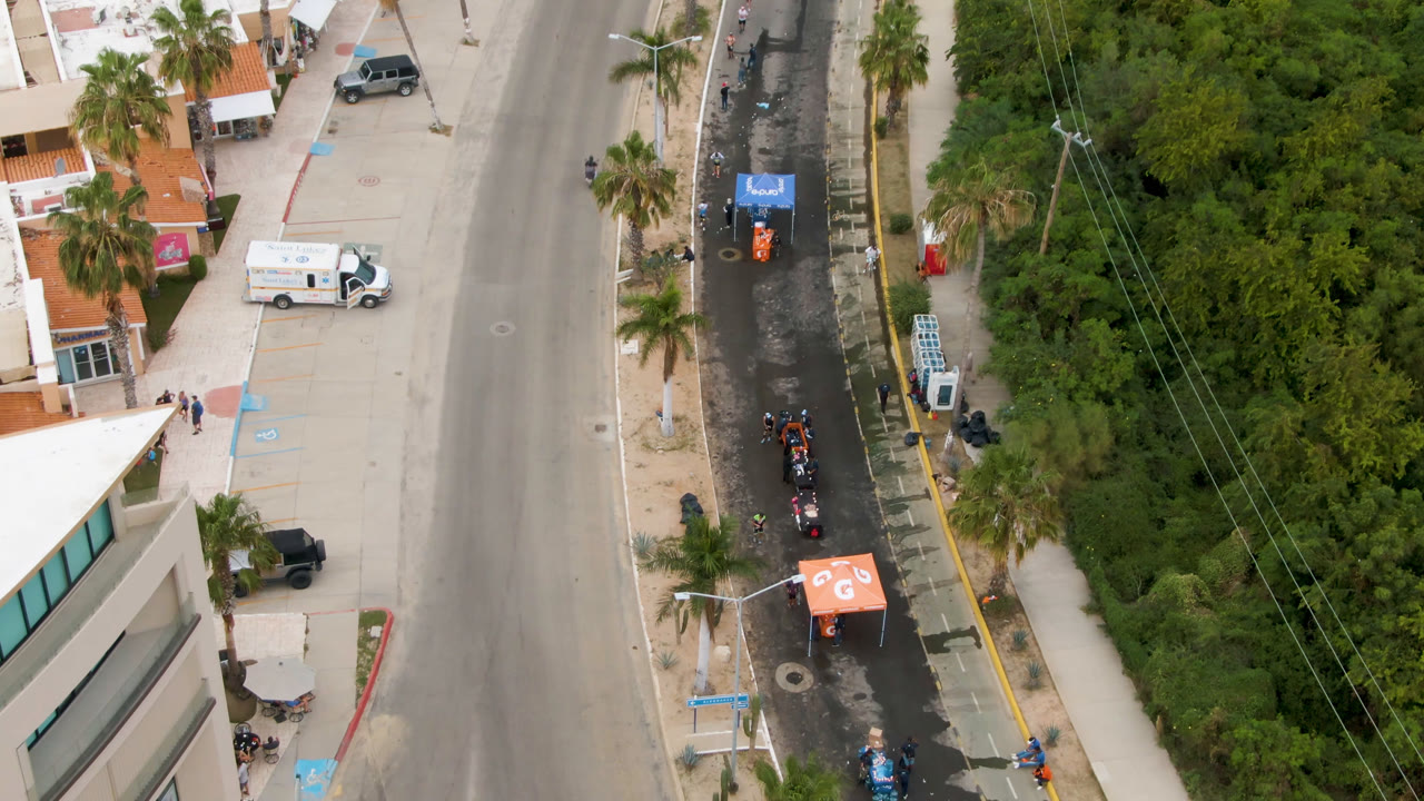 Aerial frame in San Jose Del Cabo, Mexico, with the street centered, orange and blue canopy tents along the shoulder, parked vehicles near buildings, and dense vegetation at right.
