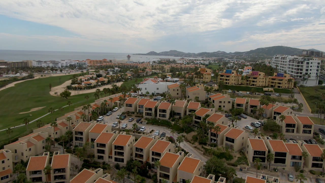 Aerial frame in San Jose del Cabo showing clustered residential buildings in front, a manicured fairway to the left, ocean on the horizon, hills in back, and no visible people.