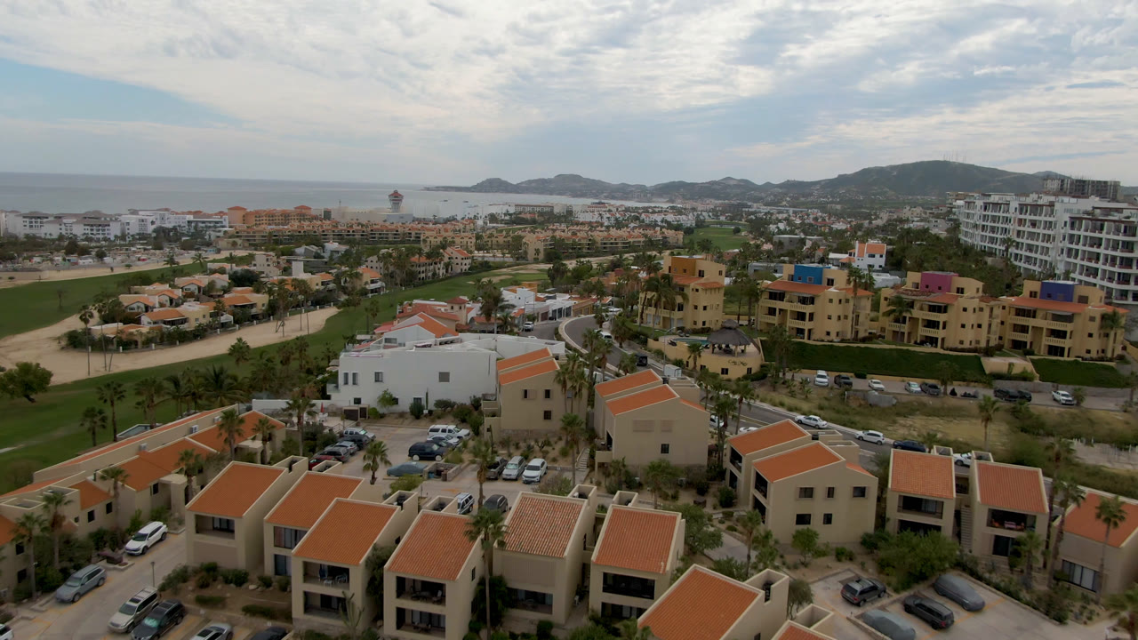 Second frame in San Jose del Cabo showing a wider coastal neighborhood, fairway greens, curved roads, distant beach and ocean, hills, clouds, and no visible people.