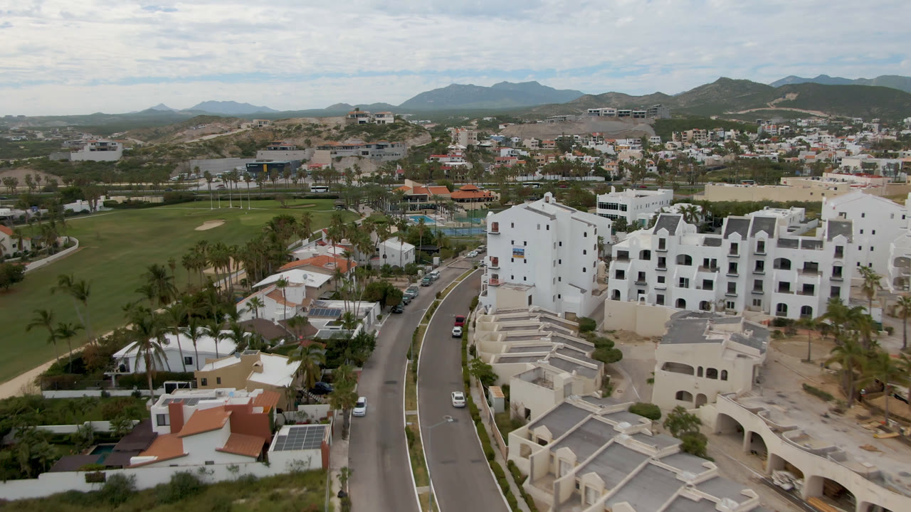 Drone-height view in San Jose del Cabo with white buildings near a curved road, small cars, palms, golf fairways, scattered homes, distant hills, and no visible people.