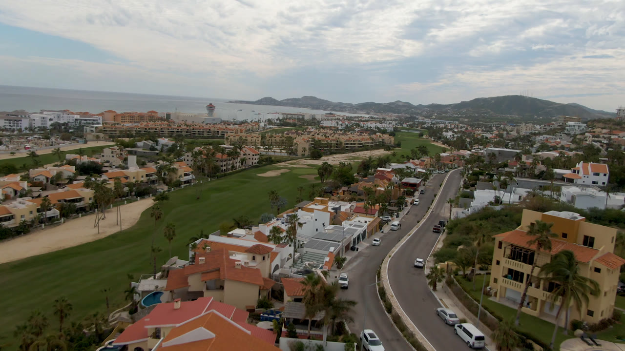 Opening aerial frame over San Jose del Cabo with green golf fairways, terracotta and white homes, a curving road, distant hills, ocean, cloud cover, and no visible people.