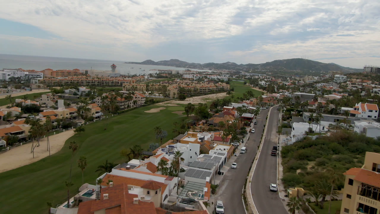 Aerial frame in San Jose del Cabo looking over a road running between homes and a broad green golf course, with ocean and low hills under a cloudy sky and no visible people.