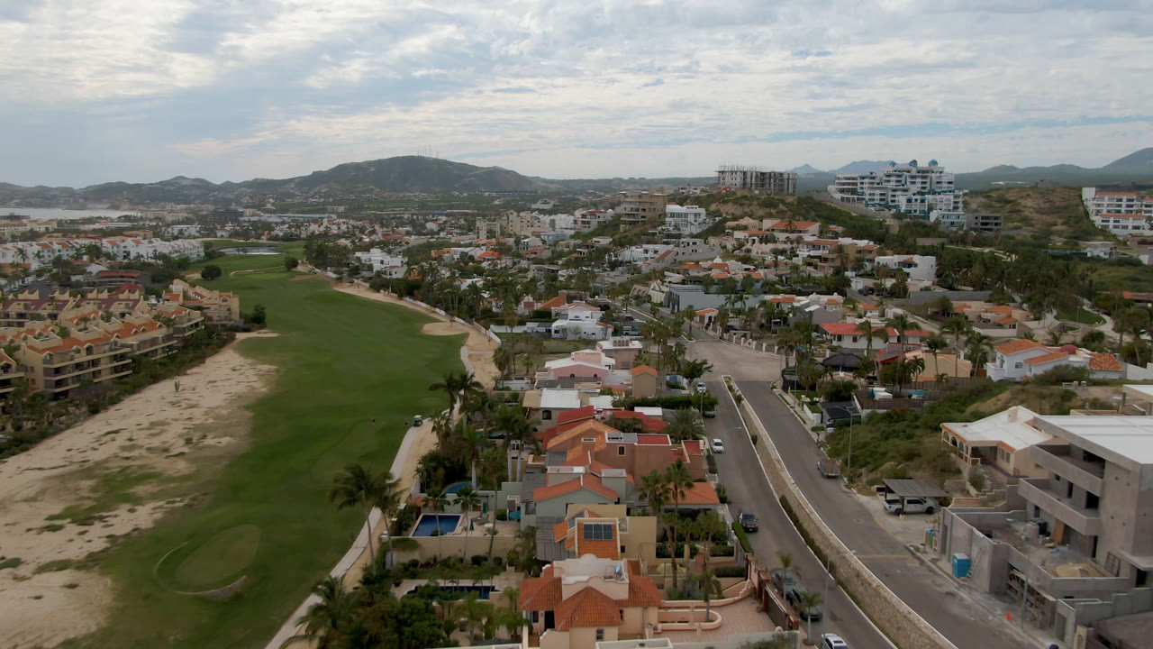Opening aerial frame over San Jose del Cabo with a green golf fairway at left, residential homes and roads at center, distant hills under cloud cover, and no visible people.
