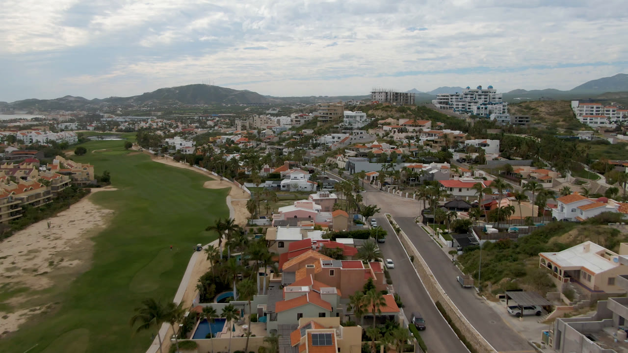 Aerial view in San Jose del Cabo looking past red-tile roofs and a straight road toward golf greens, hillside buildings, low mountains, and no visible people.