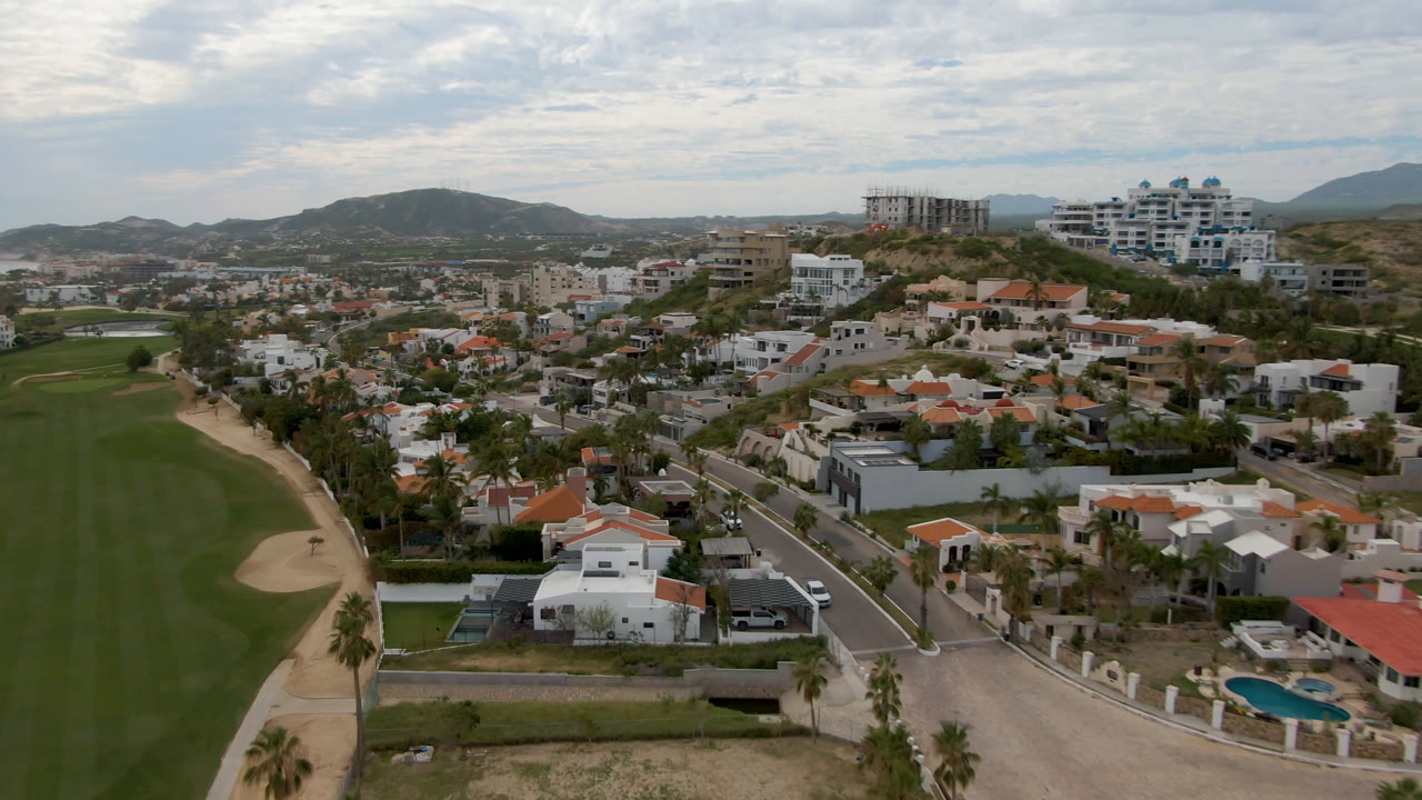 Aerial view in San Jose del Cabo with the golf fairway centered, a pale sand bunker at left, villas and larger buildings beyond, distant water, and no visible people.