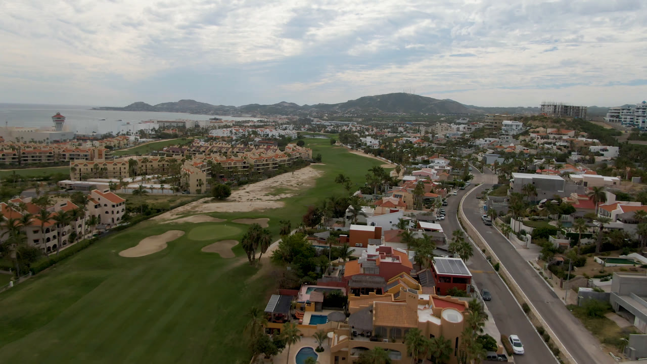 Aerial frame over San Jose del Cabo showing green fairways, sand traps, a curving road, red-roofed homes, ocean water, distant hills, and no visible people.