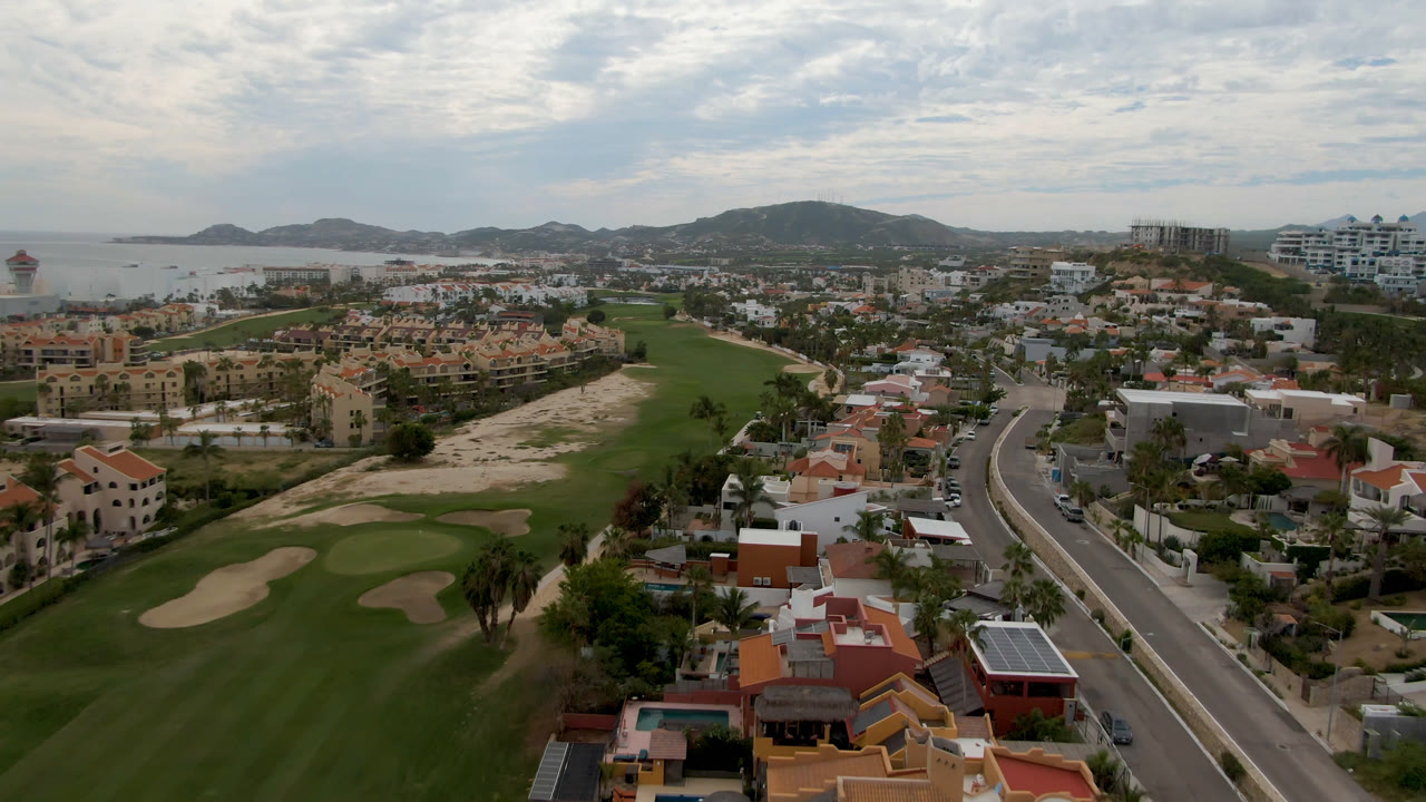 San Jose del Cabo aerial still with manicured fairways at left, a road through the center, residential blocks on both sides, coastal water, hills, and no visible people.