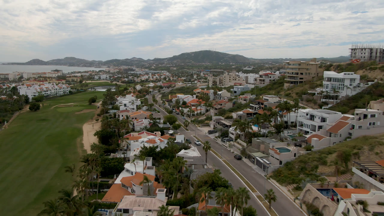 Aerial frame over San Jose del Cabo showing a green golf fairway at left, white homes and palm-lined streets below, distant hills, ocean water, cloudy sky, and no visible people.