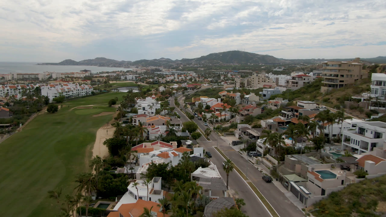 Aerial view in San Jose del Cabo with the golf fairway running through the lower left, a road and villas in the center, hillside buildings to the right, ocean on the horizon, and no visible people.