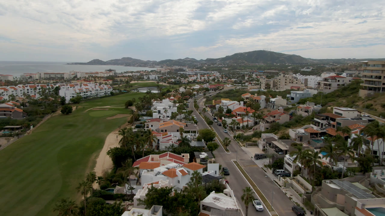 Drone view of golf course, villas, road, hills, and ocean under broken clouds.