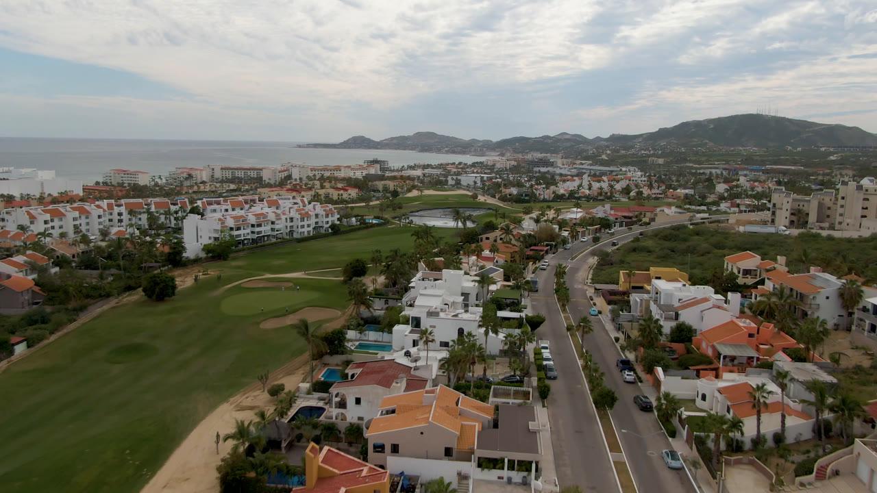 Aerial view over a golf course, road, homes, ocean, and hills.
