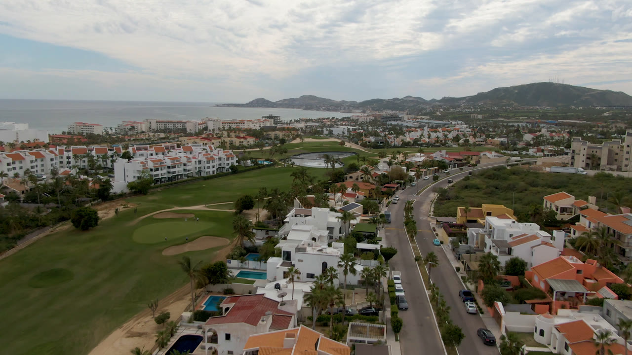 Drone view in San Jose del Cabo with sand bunkers and fairways in front, coastal housing and a road through the middle, ocean and hills beyond, and no visible people.
