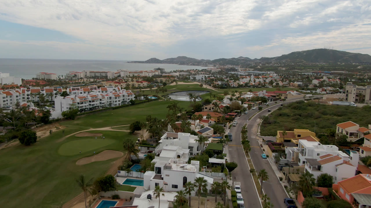 Aerial frame in San Jose del Cabo showing a wide golf green, nearby white buildings, a divided road, distant ocean water, hillside terrain, and no visible people.