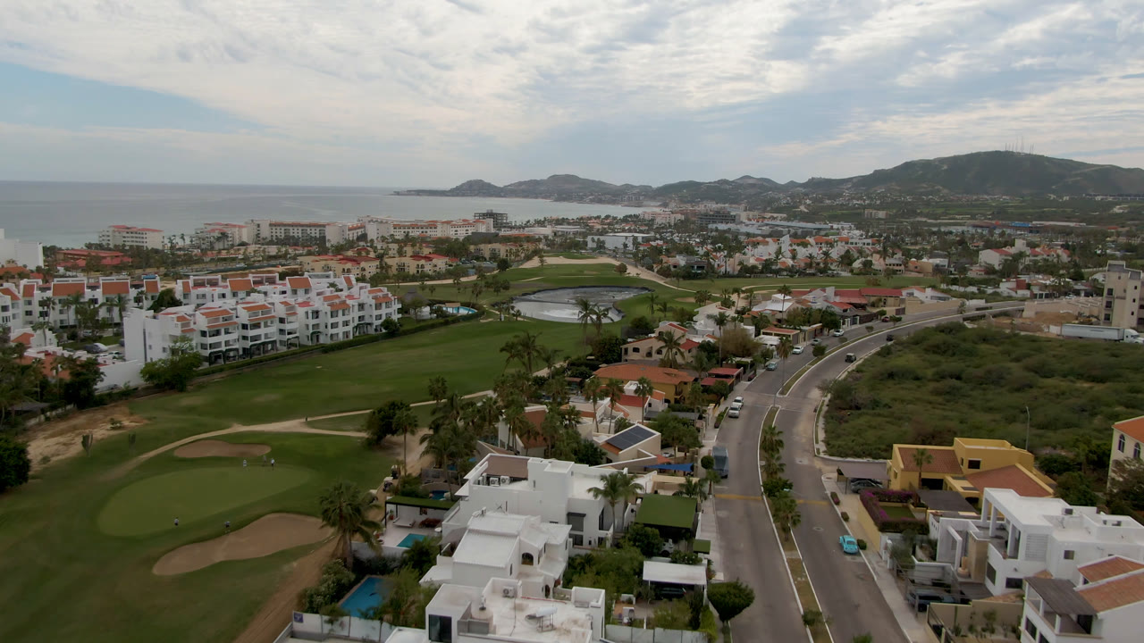 Wide aerial of golf greens, resort-style homes, roadway, ocean, and hills.