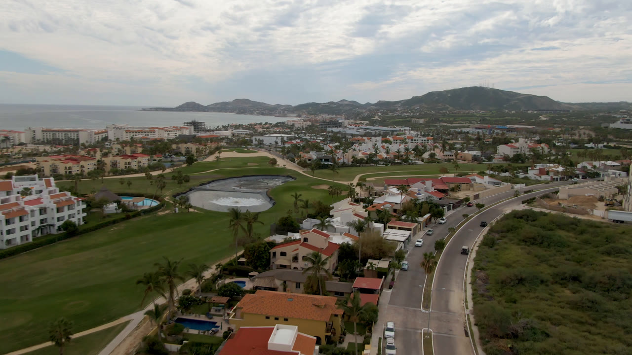 Aerial San Jose del Cabo frame looking across a dark golf course water hazard, nearby houses, winding roads, ocean edge, low hills, and no visible people.