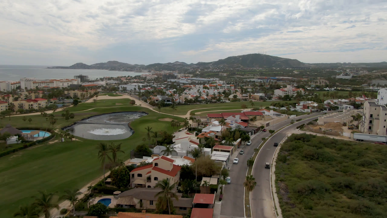 Wide drone view over San Jose del Cabo showing a curving divided road in the foreground, golf course greens and water, clustered homes, coastline, hills, and no visible people.