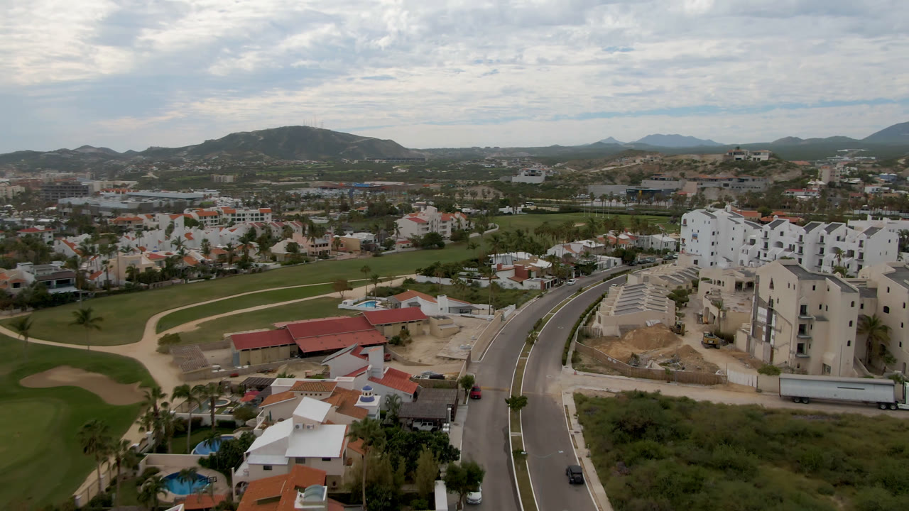 Aerial frame over San Jose del Cabo showing a curving road, golf course grass, white residential buildings, red roofs, distant hills, and no visible people.