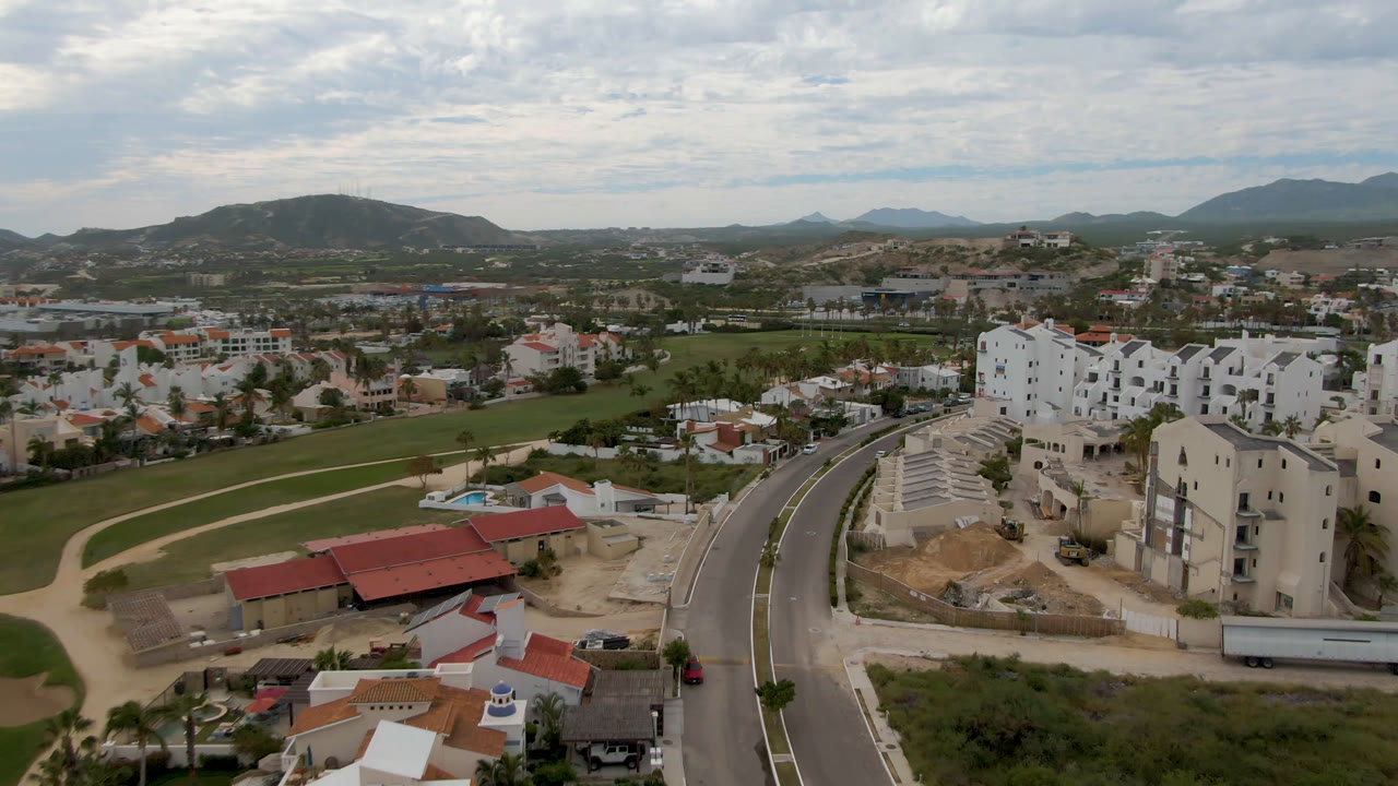 Drone view in San Jose del Cabo looking over green fairways, scattered homes, white buildings, low mountains, and a cloudy sky, with no visible people.