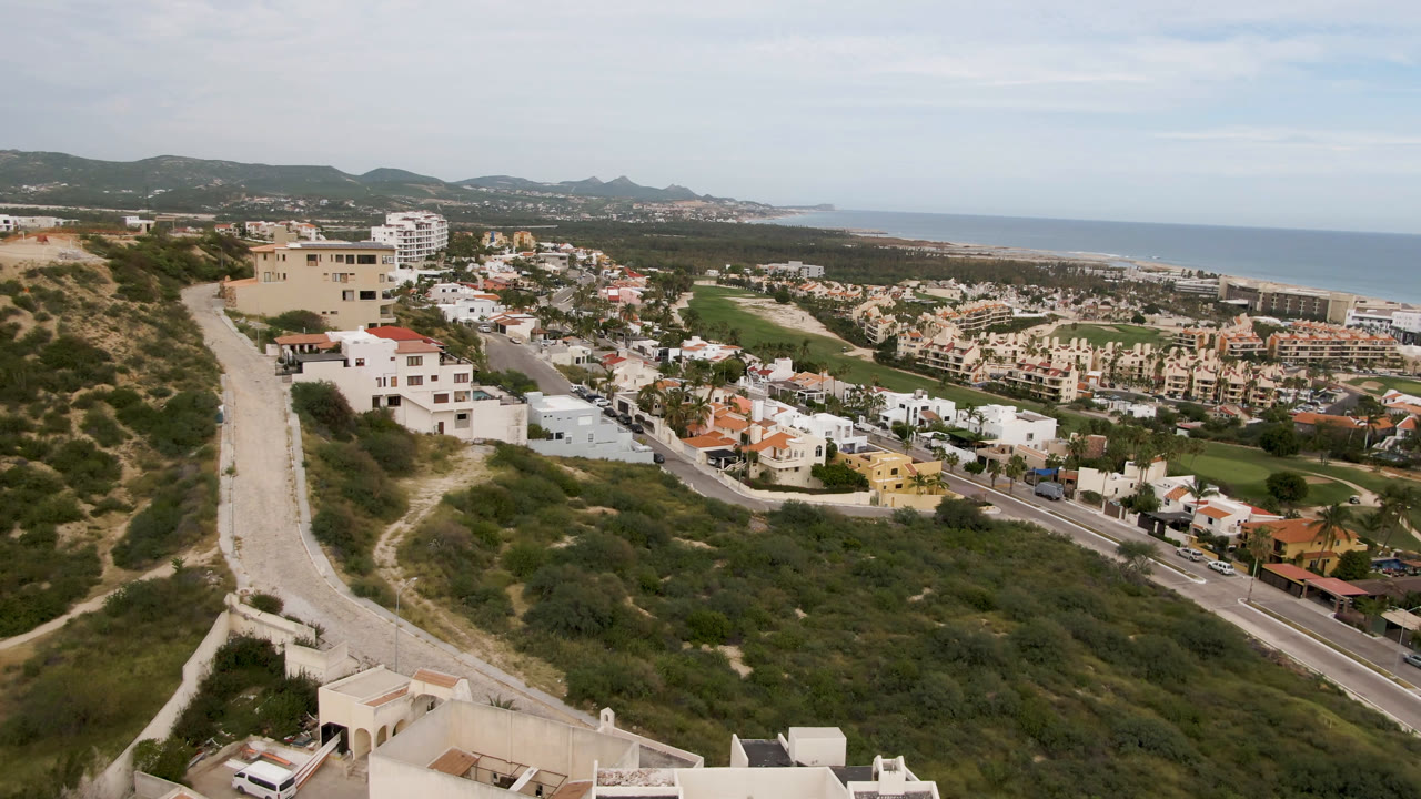 Opening aerial frame of San Jose del Cabo with scrub-covered hillside in front, white homes and a road below, resort buildings and ocean beyond, and no visible people.
