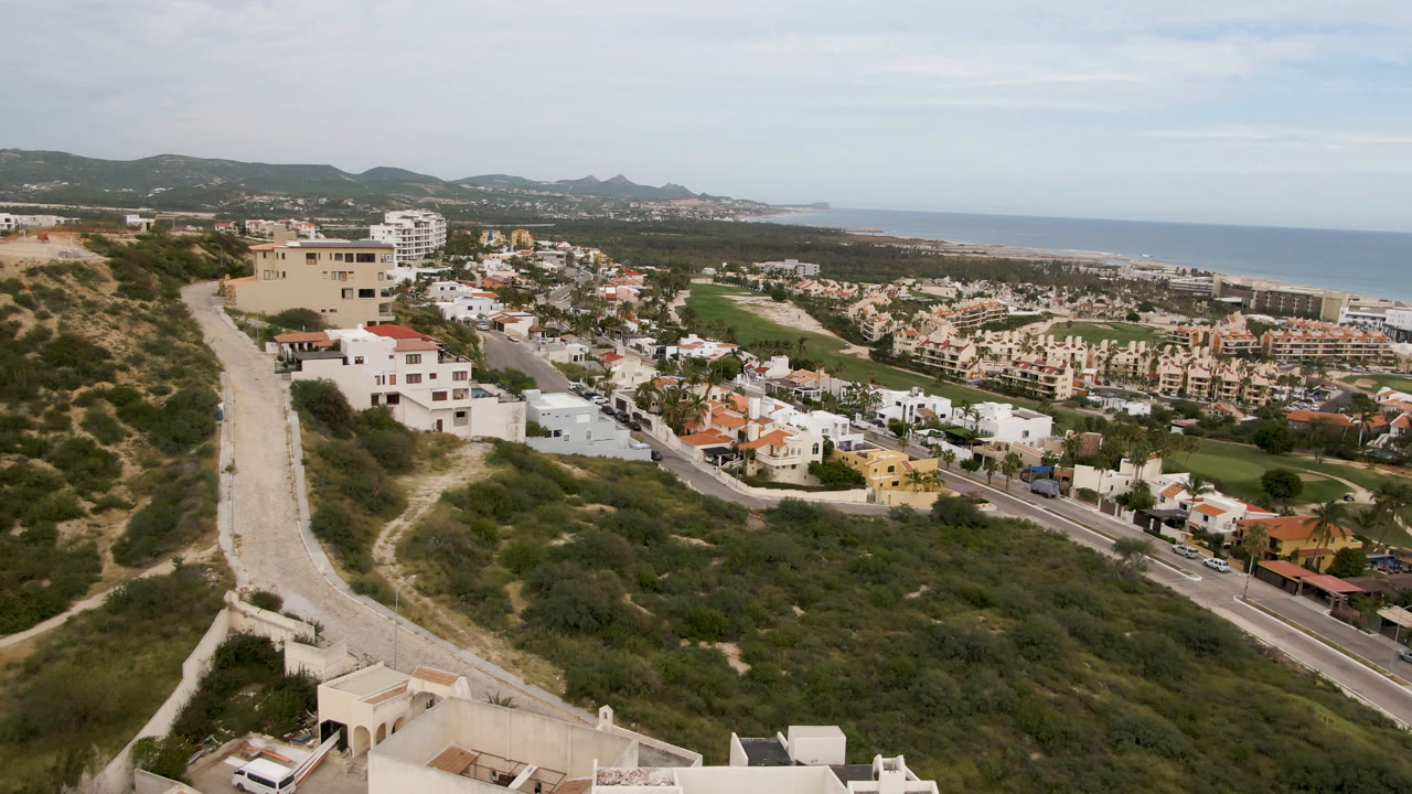 Second aerial frame of San Jose del Cabo showing more of the hillside road at left, dense homes in the center, green fairway areas near the coast, ocean on the horizon, and no visible people.