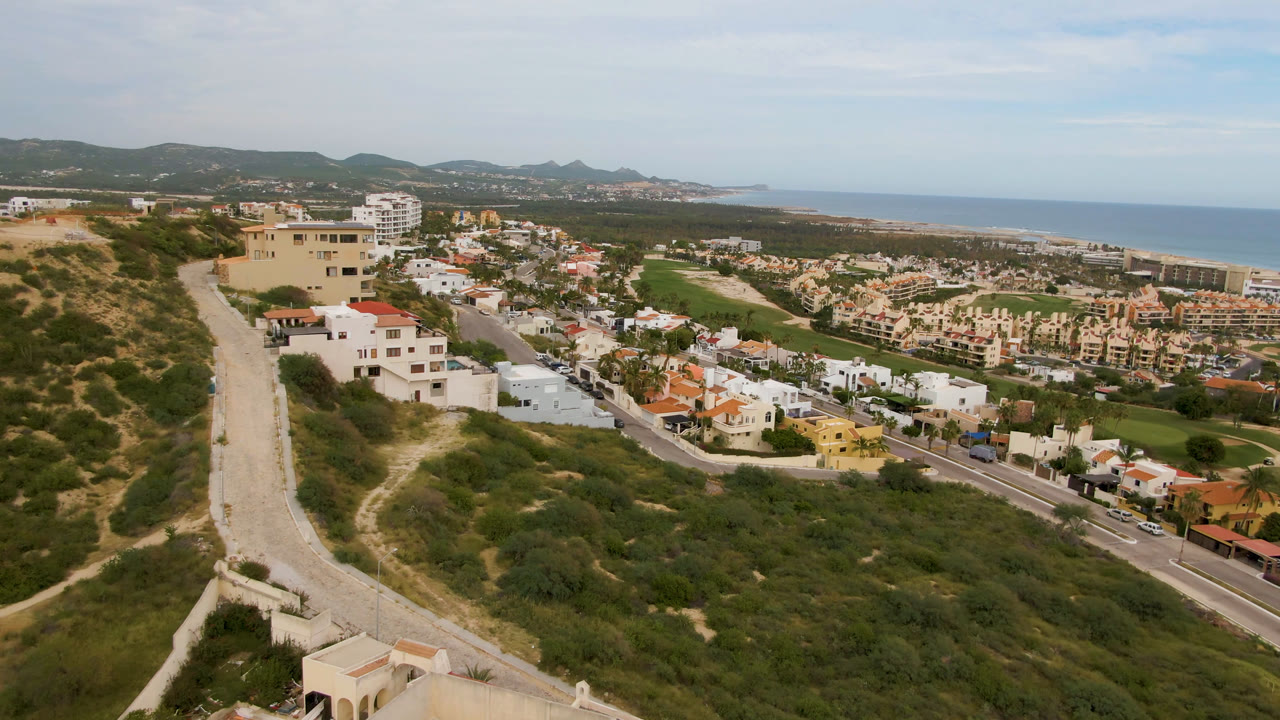 Aerial footage over San Jose del Cabo frames the left hillside road, clustered residential buildings, a green golf-course area, distant coast, blue ocean, and no visible people.
