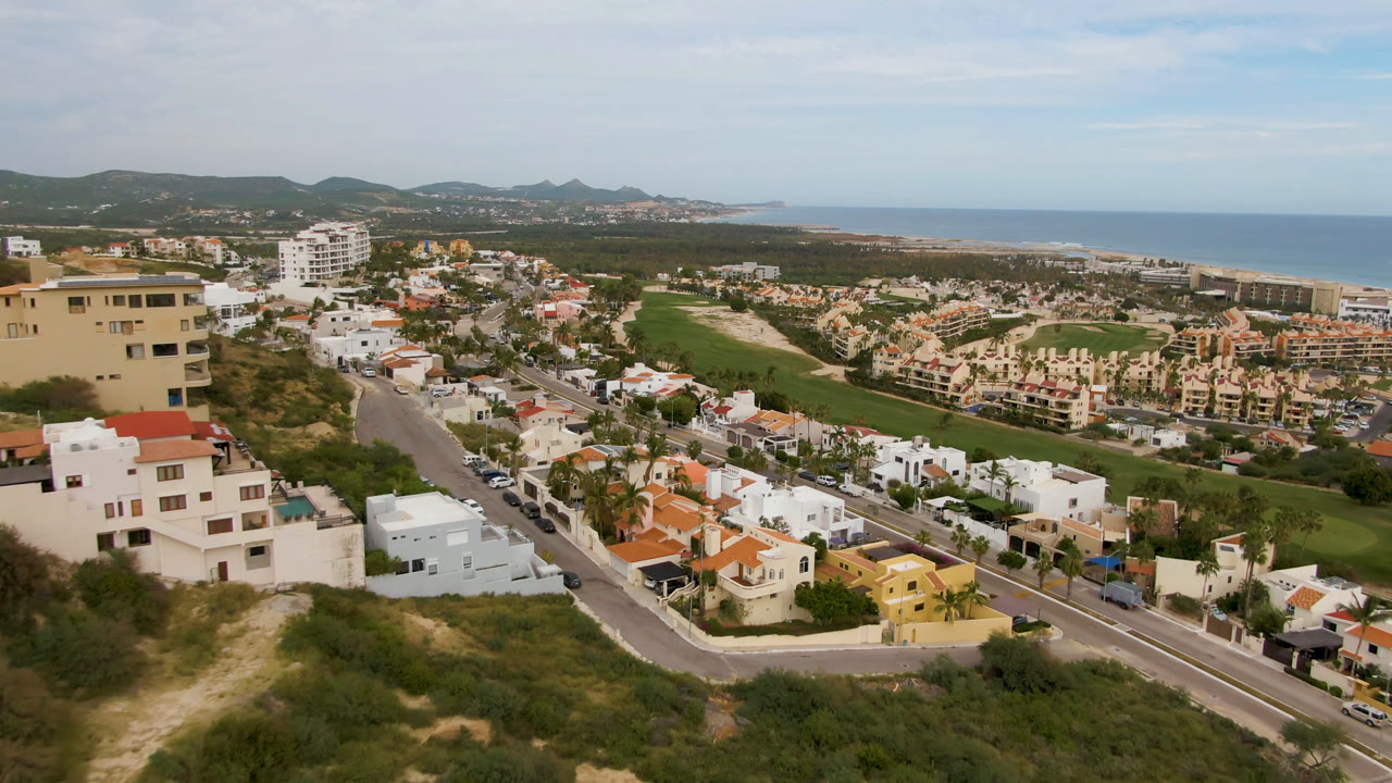 Aerial frame in San Jose Del Cabo showing a broad residential area, a green golf fairway through the middle distance, ocean on the right, low hills beyond, and no visible people.