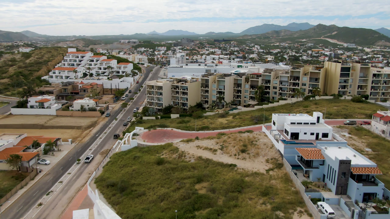 Drone view of a road, apartment buildings, open lots, hills, and distant mountains.