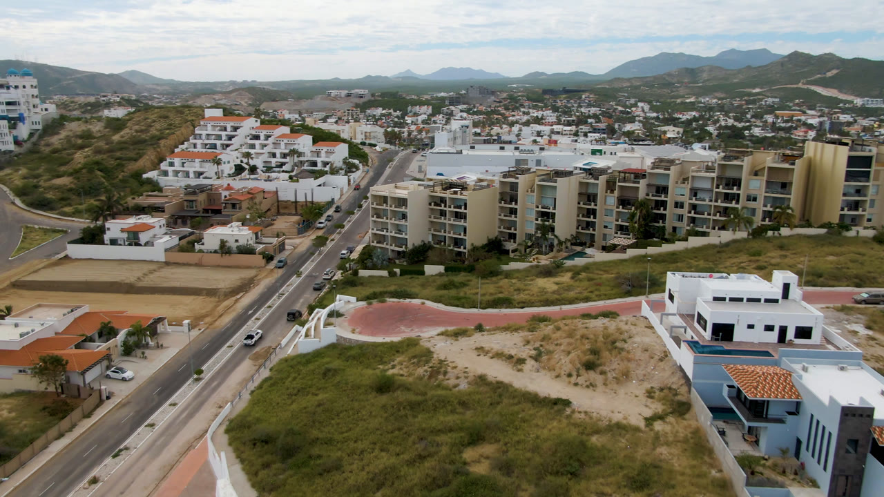 Second aerial frame in San Jose del Cabo looking across a curving hillside road, stacked white residences, beige apartment blocks, undeveloped lots, distant city blocks, and no visible people.