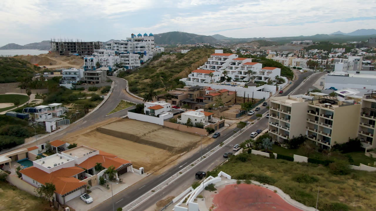 Aerial frame in San Jose del Cabo showing white residential blocks, a broad road, parked cars, a tan open field, hills, distant water, and no visible people.