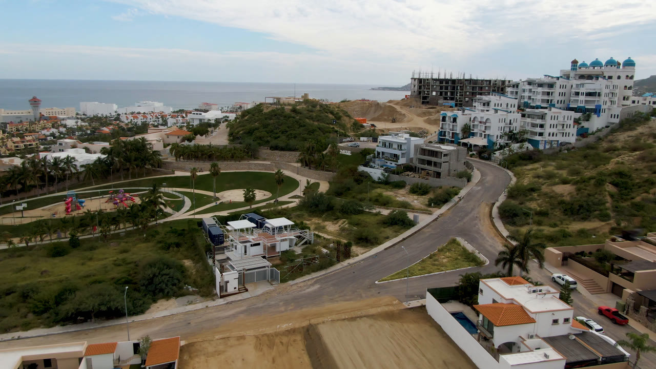 Aerial frame from San Jose del Cabo showing a curved road below hillside homes, an unfinished building on the ridge, the ocean horizon, and no visible people.