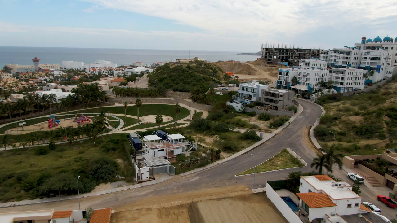 Aerial view in San Jose del Cabo with a landscaped circular park in the midground, white hillside buildings, a winding road, the sea beyond, and no visible people.