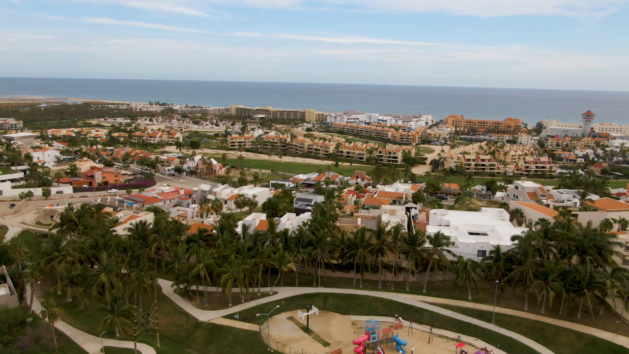 Wide aerial view of coastal buildings, palms, paths, beach, ocean, and sky.