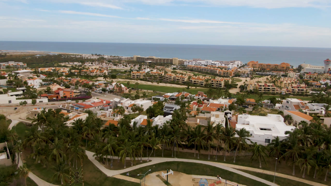 One second into the San Jose del Cabo aerial, the frame holds white-roofed buildings, palms, resort blocks, a distant strip of beach, blue ocean, and no visible people.