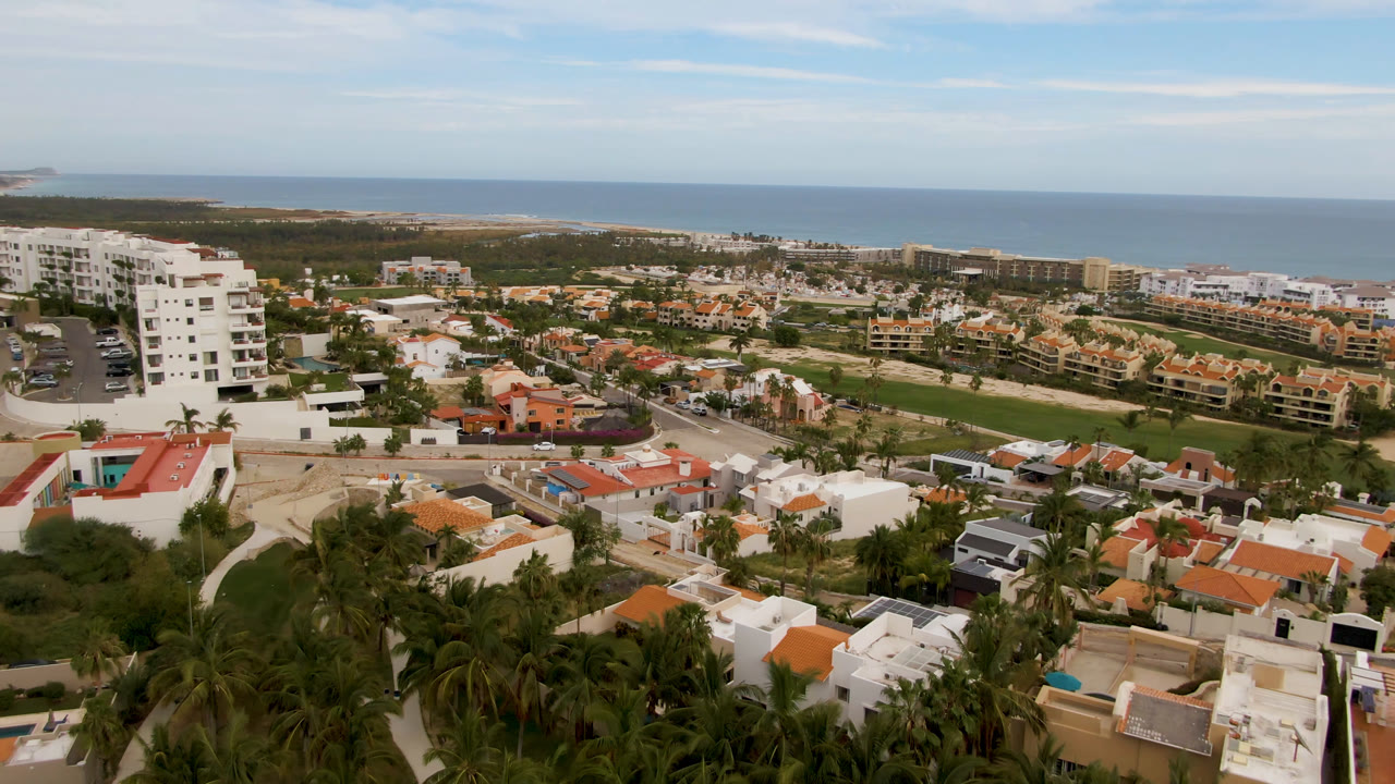 Elevated San Jose del Cabo view with a white apartment block at left, low homes across the center, ocean in the distance, and no visible people.