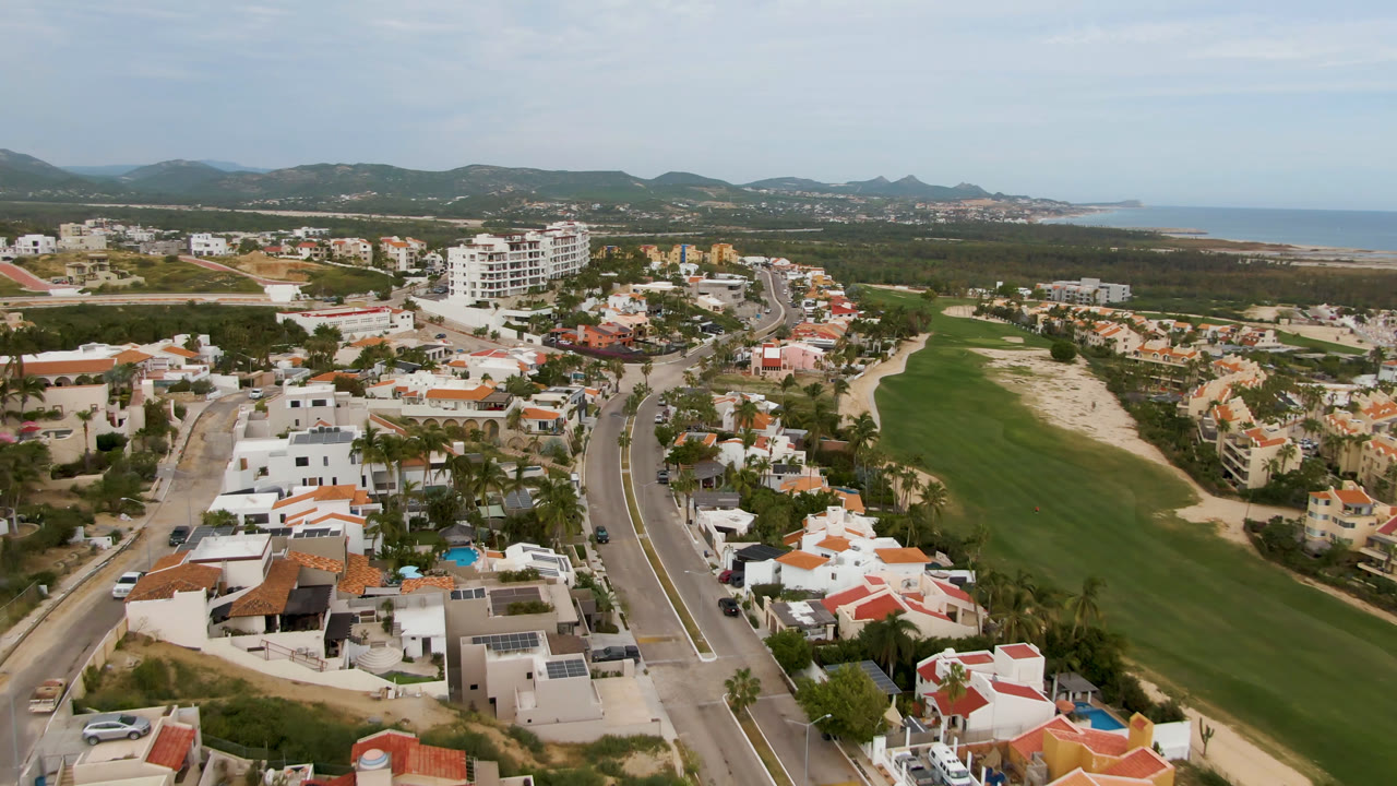 High aerial view over San Jose del Cabo with white homes, terracotta roofs, a curving road, a green golf fairway, distant hills, and no visible people.