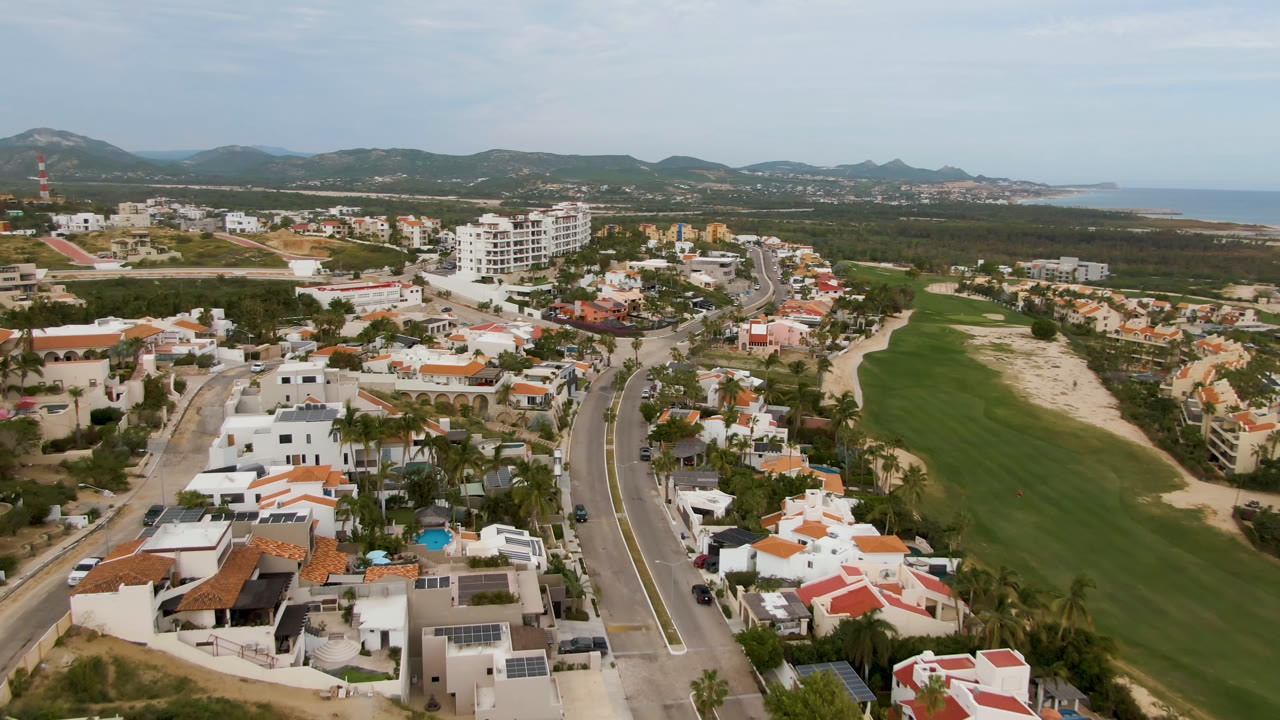 Aerial frame in San Jose del Cabo showing residential roofs and palms in the foreground, a winding road, a broad golf fairway, distant buildings, hills, and no visible people.