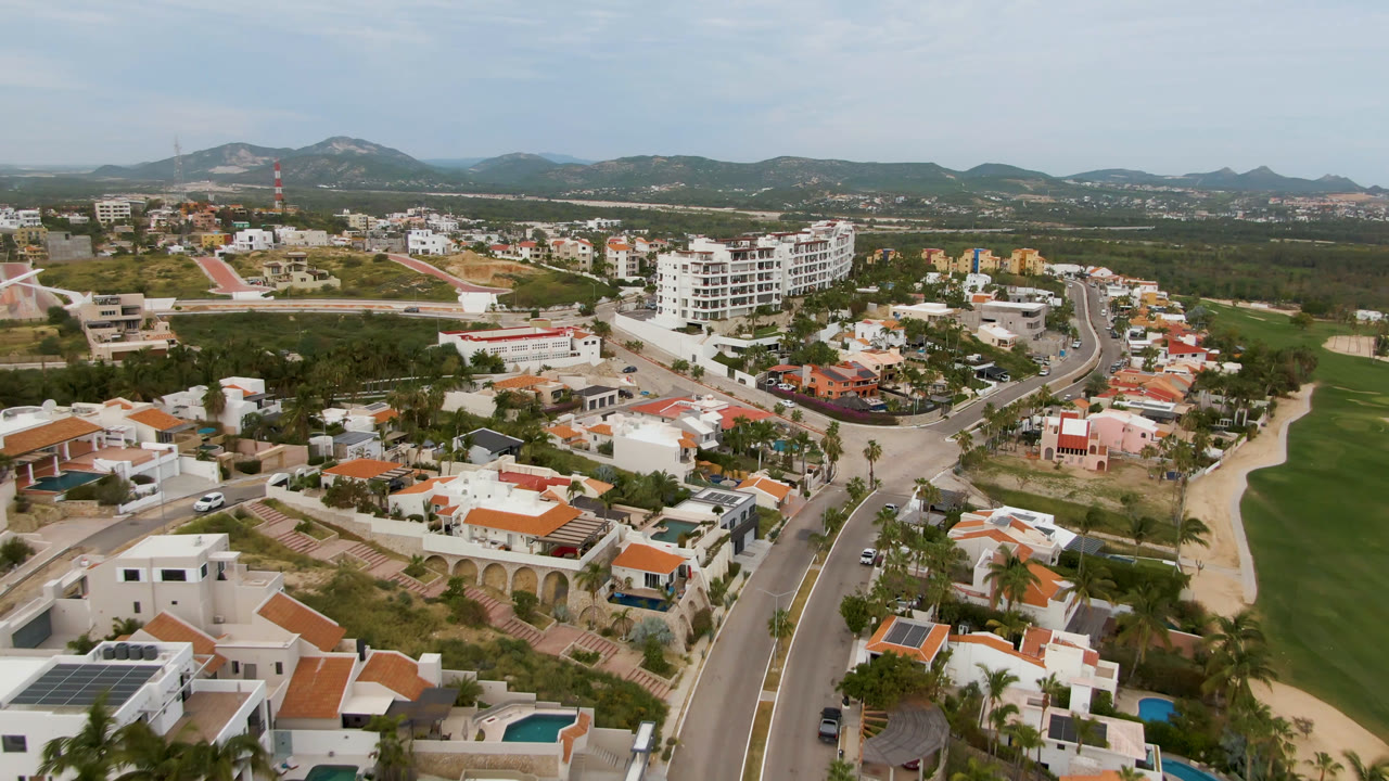 San Jose del Cabo aerial frame showing a wider road junction, clustered homes, a green open area, mid-rise buildings, hills, and no visible people.