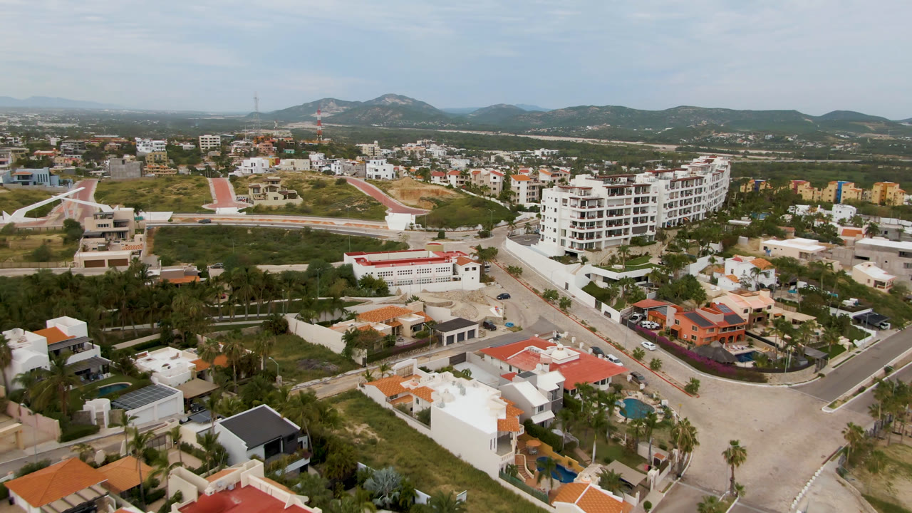 Aerial frame in San Jose del Cabo looking past houses and a red-roofed low building toward a white apartment block, hillside greenery, distant hills, and no visible people.