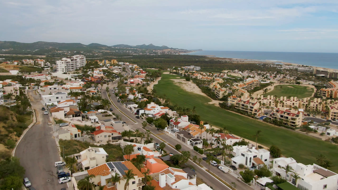 Drone view in San Jose del Cabo looking across a paved road and clustered homes toward a green golf course, low hills, blue ocean, and no visible people.