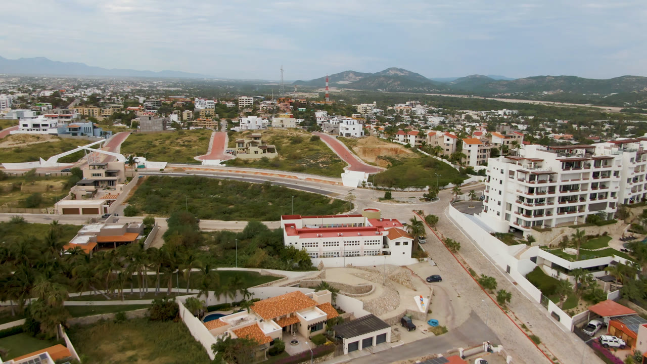 Aerial view over San Jose del Cabo with a curving road, white residential buildings, red roofs, green hillside, distant mountains, and no visible people.