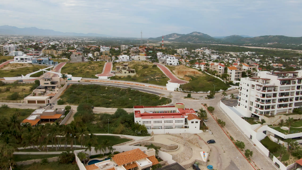 Drone-style aerial over San Jose del Cabo showing a red-roofed building, a winding road across the hillside, clustered homes, hazy mountains, and no visible people.