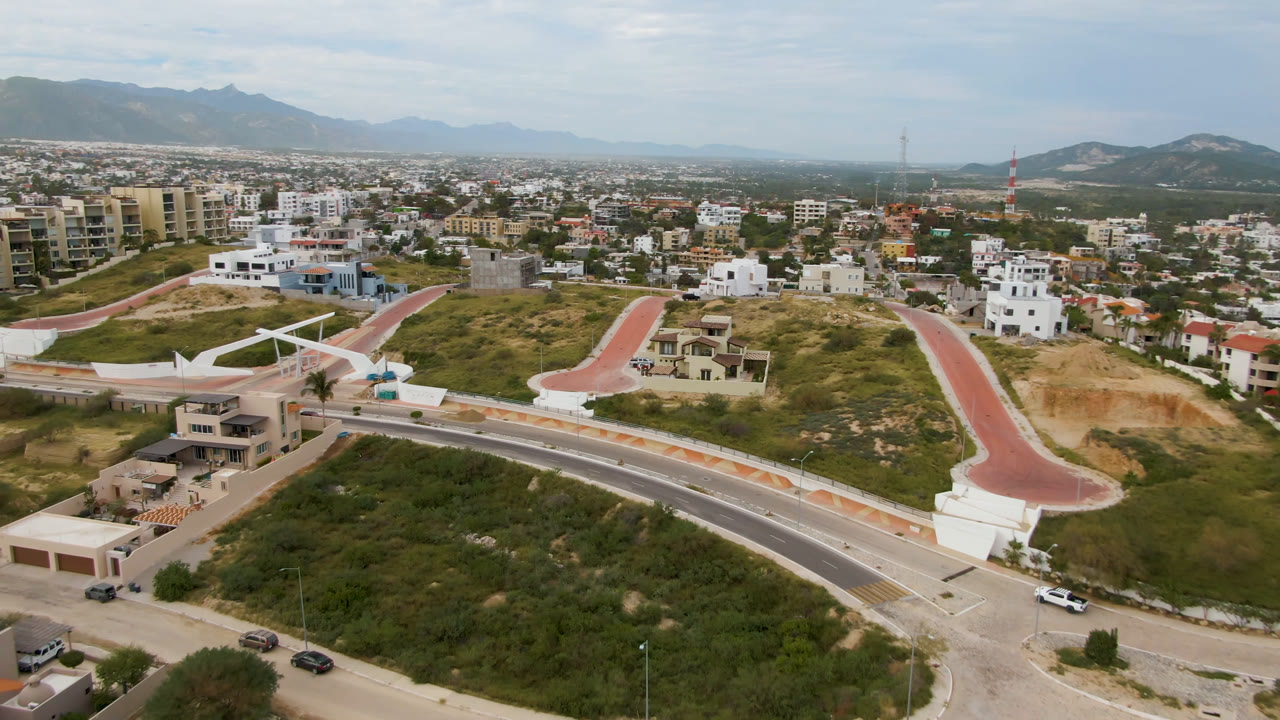 Aerial frame in San Jose del Cabo showing a curved roadway below, a red bridge-like paved route across the hillside, modern residential buildings, vehicles, mountains, and no visible people.