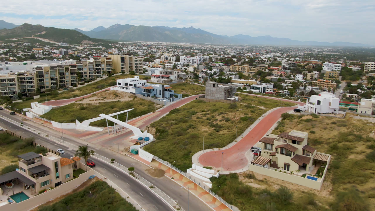 Drone view in San Jose del Cabo showing a curving red paved path beside vacant lots, nearby apartment buildings, cars on the road, distant mountains, and no visible people.